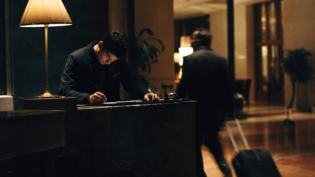 Night receptionist at a hotel front desk writing a note while a guest walks past with luggage