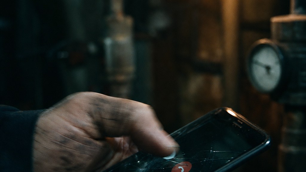 Close crop of a grease-marked hand tapping a call button on a cracked phone screen in a boiler room