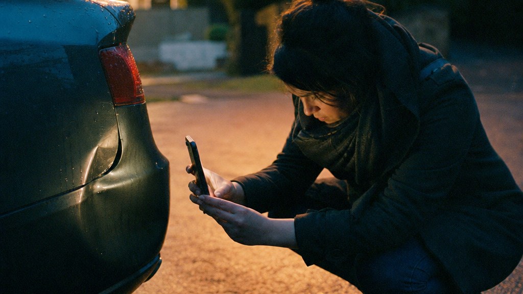 Woman on a suburban driveway at dusk photographing a dented car bumper with her phone