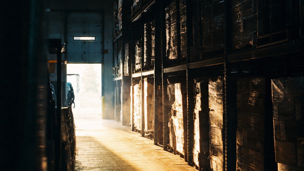 Long warehouse aisle of shrink-wrapped pallets with one empty bay and a shaft of daylight from a dock door
