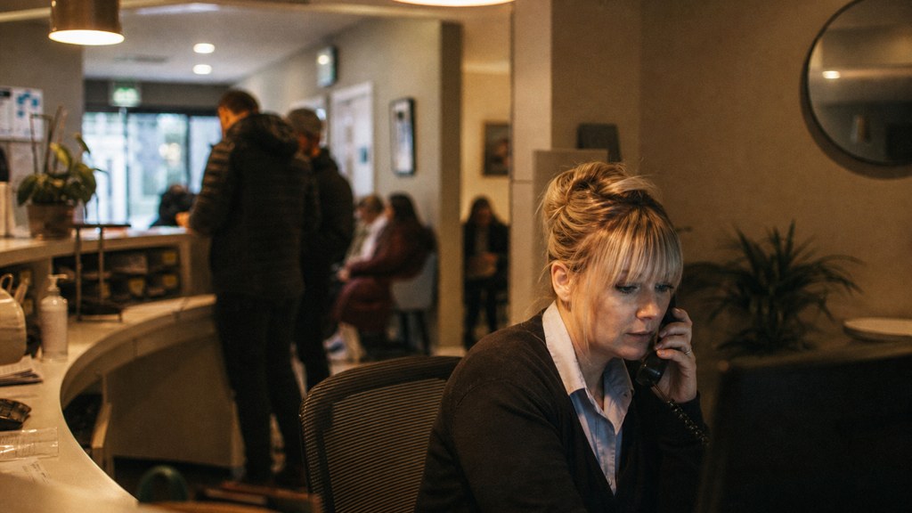 A clinic receptionist taking a phone call at a busy front desk with patients waiting, photographed in Cardiff