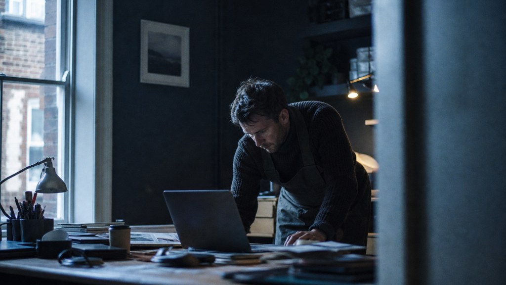 A small-business owner in an apron leaning over a laptop on a cluttered back-office desk late in the day, photographed in Dublin