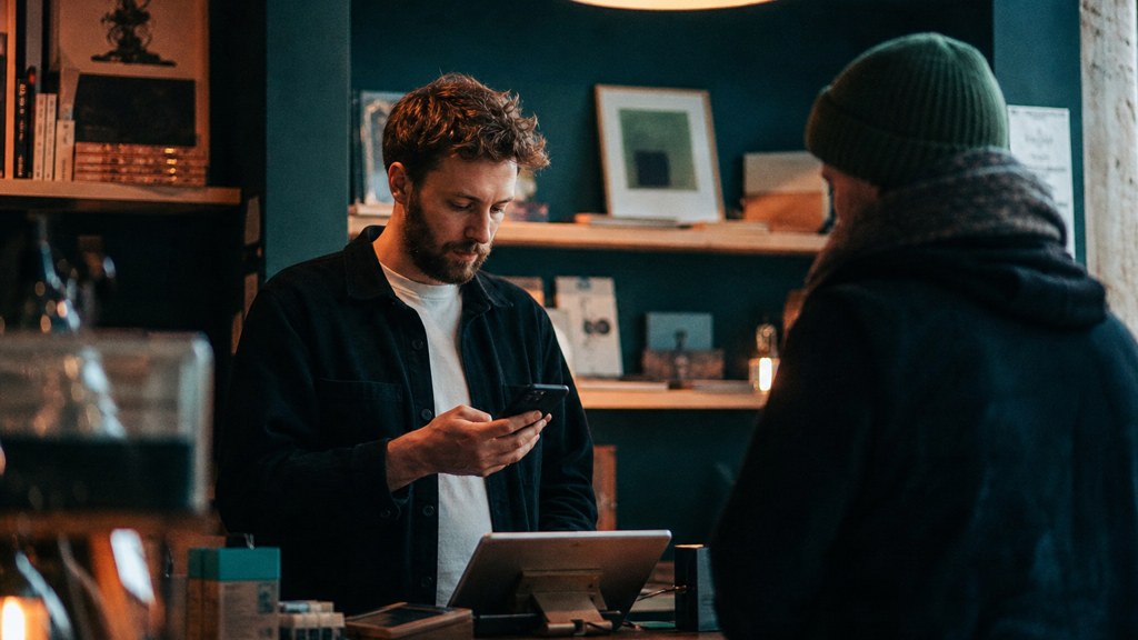 A shop manager standing behind the counter checking a phone while a customer waits at the till, photographed in Dublin