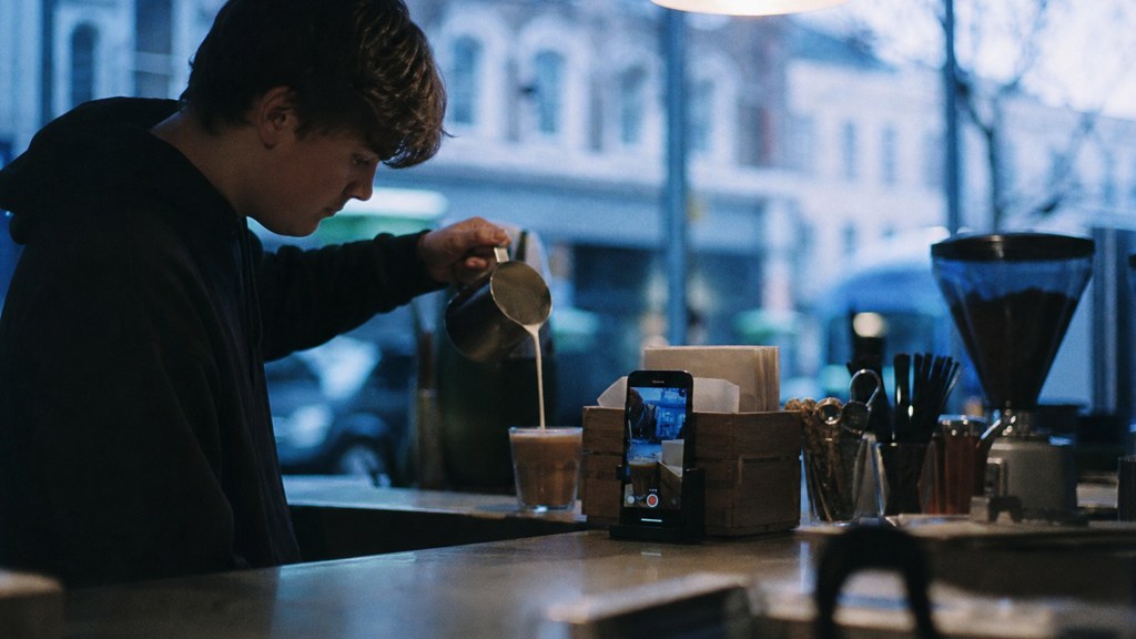 A young cafe worker filming a drink being poured on a phone propped against a napkin holder, photographed in Dublin