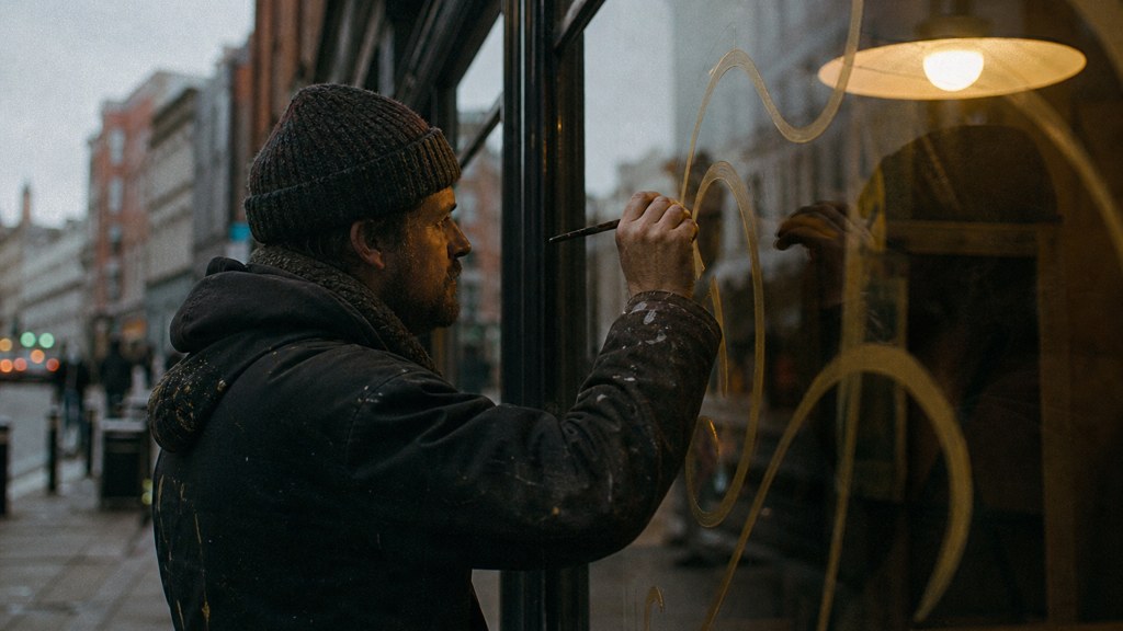 A sign painter finishing gold lettering on a shopfront window from the street side, photographed in Dublin
