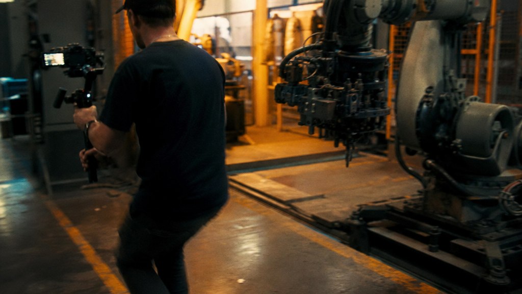 Videographer walking backwards with a gimbal ahead of a robot arm in a bright assembly cell