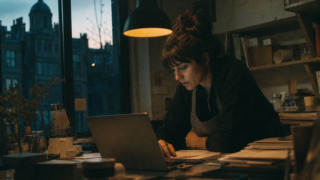 A small-business owner in an apron leaning over a laptop on a cluttered back-office desk late in the day, photographed in Edinburgh