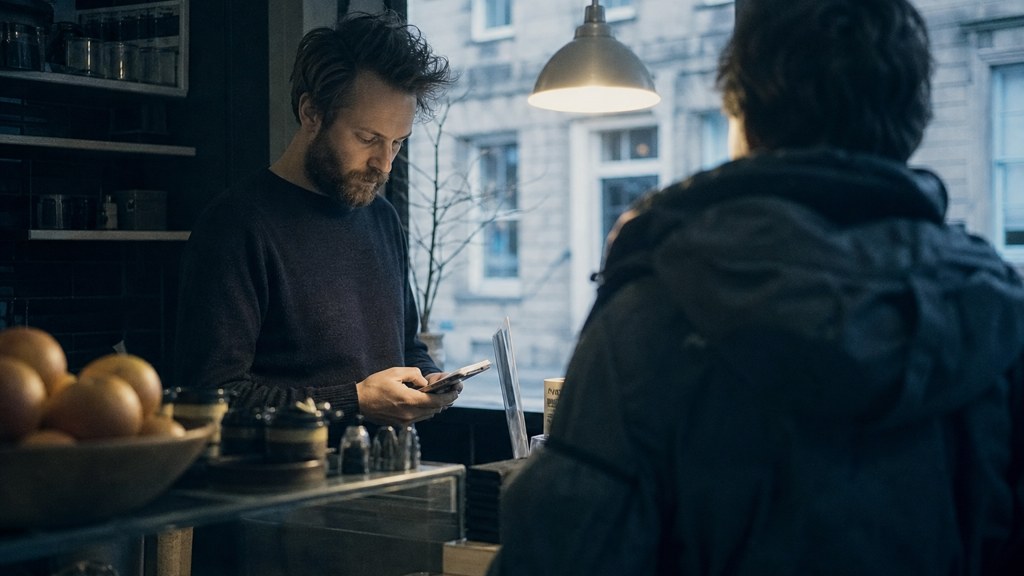 A shop manager standing behind the counter checking a phone while a customer waits at the till, photographed in Edinburgh