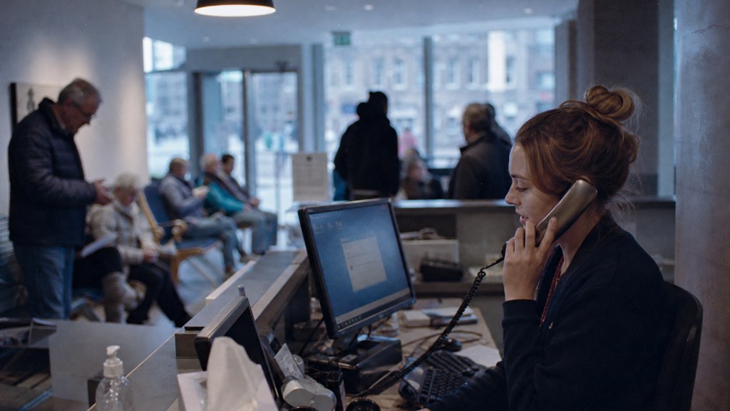 A clinic receptionist taking a phone call at a busy front desk with patients waiting, photographed in Edinburgh