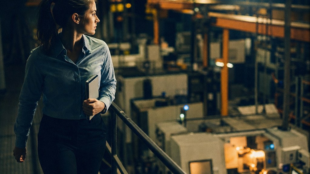 Industrial marketer on a mezzanine walkway above a CNC line holding a tablet under amber work lamps