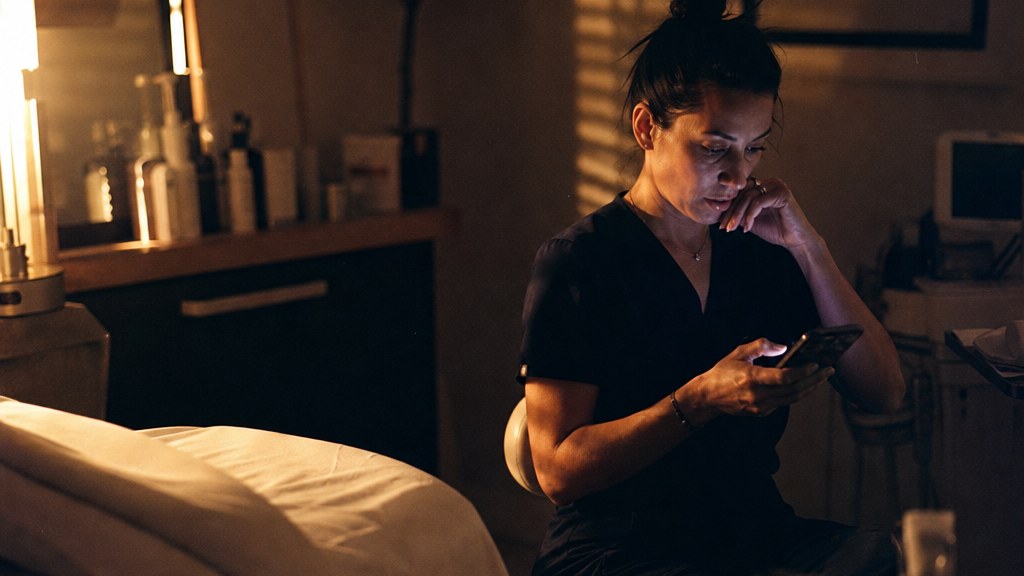 Med spa owner in scrubs on a treatment stool at night reading an AI answer on her phone with amber blinds behind her