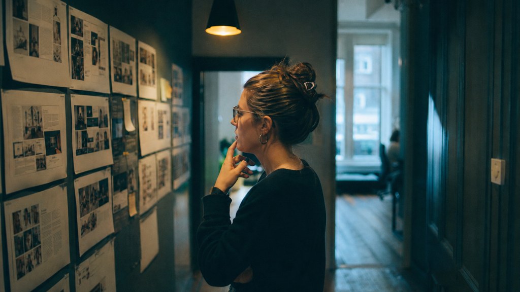 A media planner standing at a wall of printed layouts in a narrow agency corridor, photographed in Edinburgh