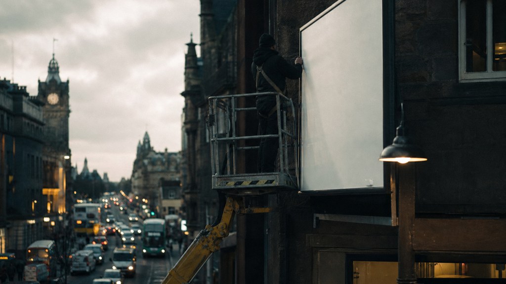 A crew member on a lift installing a blank billboard panel above a busy street, photographed in Edinburgh