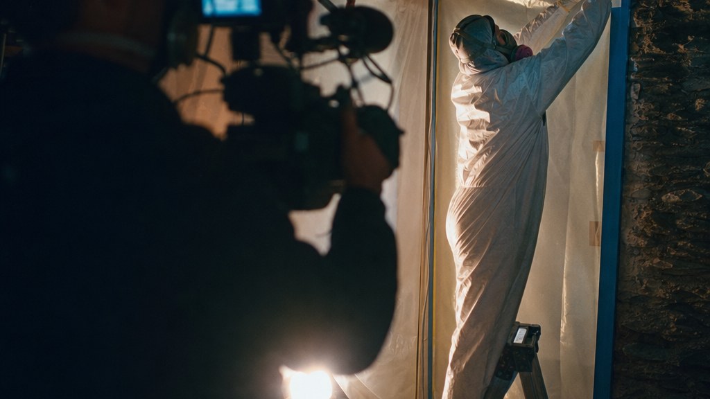 Camera operator filming a remediation technician taping plastic containment sheeting in a stripped basement under a work lamp