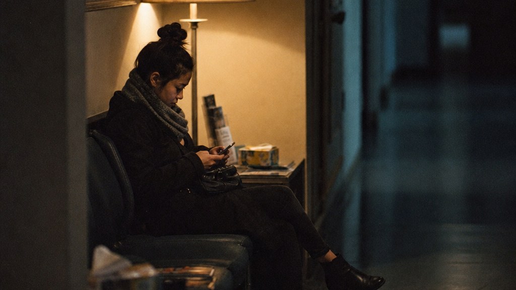 Woman sitting on a clinic waiting-room bench scrolling a treatment page on her phone with warm lamplight behind her