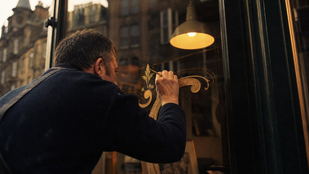 A sign painter finishing gold lettering on a shopfront window from the street side, photographed in Edinburgh