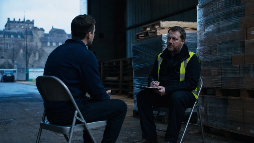 A warehouse supervisor interviewing a candidate on folding chairs beside stacked pallets, photographed in Edinburgh