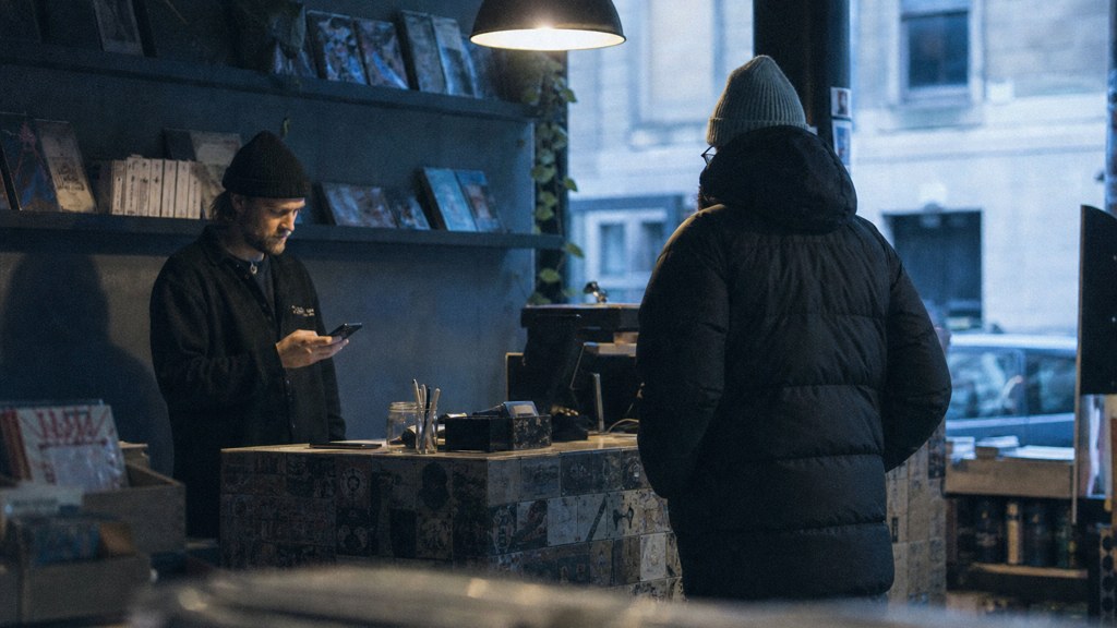 A shop manager standing behind the counter checking a phone while a customer waits at the till, photographed in Glasgow
