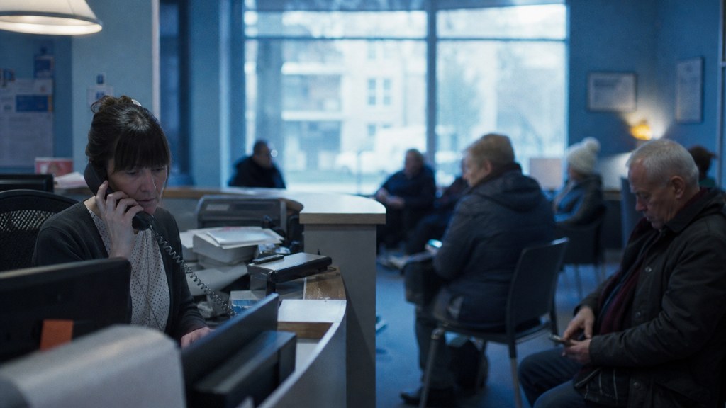 A clinic receptionist taking a phone call at a busy front desk with patients waiting, photographed in Glasgow