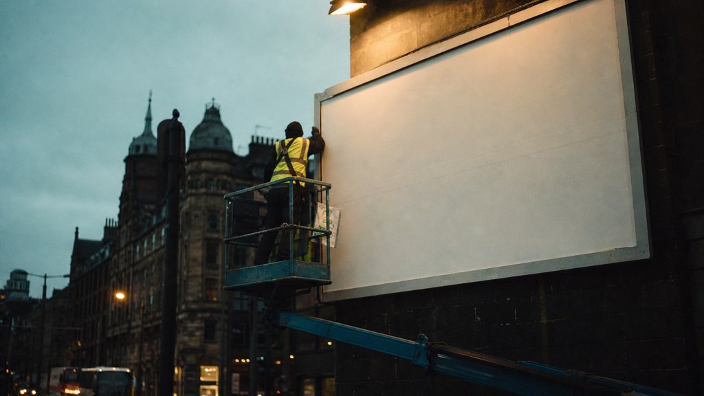 A crew member on a lift installing a blank billboard panel above a busy street, photographed in Glasgow