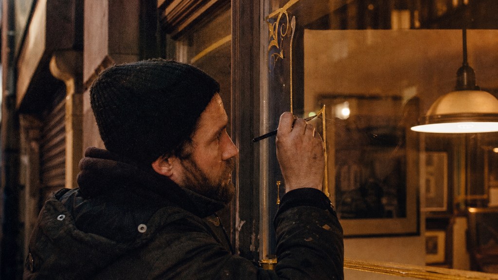 A sign painter finishing gold lettering on a shopfront window from the street side, photographed in Glasgow