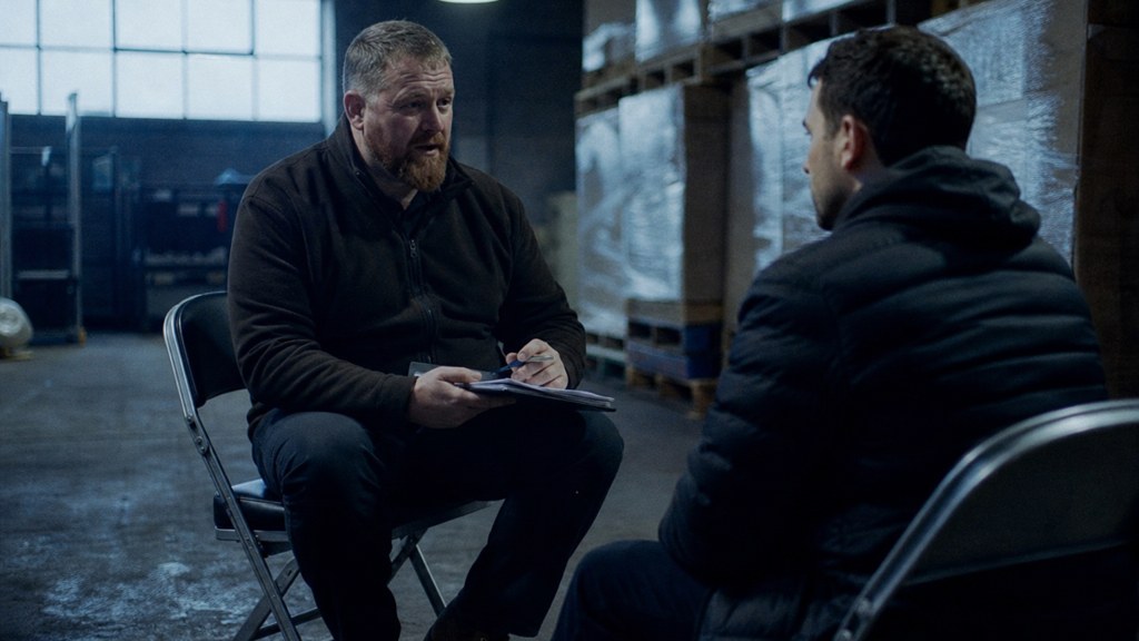 A warehouse supervisor interviewing a candidate on folding chairs beside stacked pallets, photographed in Glasgow