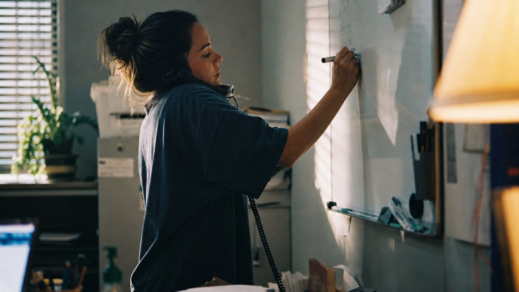 Dispatcher at a small restoration firm writing a job address on a wall whiteboard while holding a phone