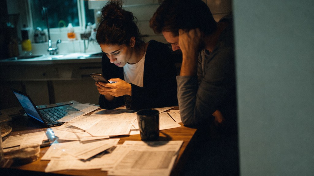 Young couple at a kitchen table at night typing a question into a phone with mortgage paperwork spread out
