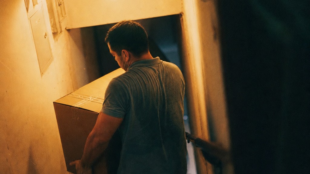 Mover carrying a wardrobe box down a narrow stairwell, caught mid-stride under a bare bulb