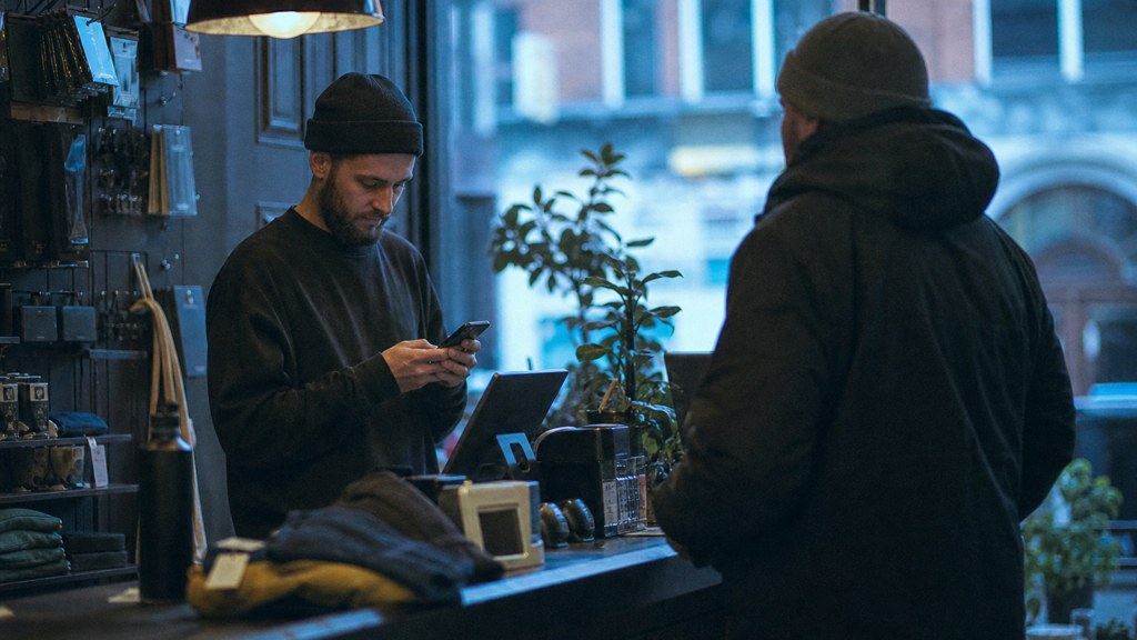 A shop manager standing behind the counter checking a phone while a customer waits at the till, photographed in Liverpool