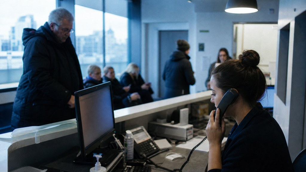 A clinic receptionist taking a phone call at a busy front desk with patients waiting, photographed in Liverpool