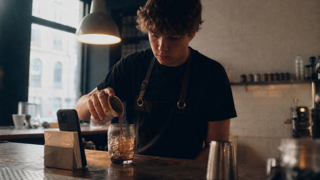 A young cafe worker filming a drink being poured on a phone propped against a napkin holder, photographed in Liverpool