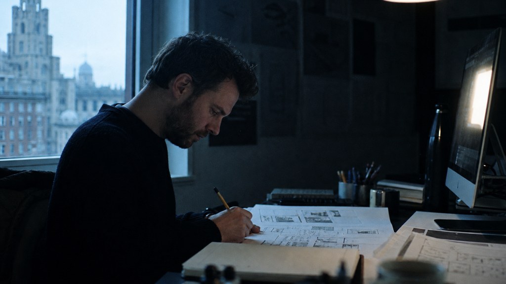 A studio designer sketching a page layout on paper beside a monitor in a dim room, photographed in Liverpool