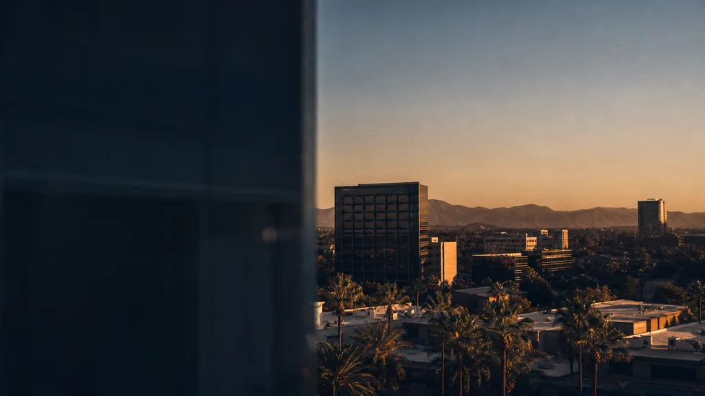 Editorial photograph of an Orange County California business district at golden hour seen past the dark edge of an office tower, with mid-rise office buildings, palm trees, low rooftops and the Santa Ana Mountains under a clear warm sky, and no people, text, signage or logos