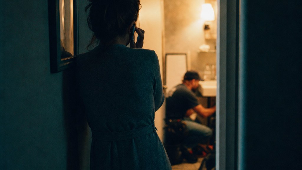 Homeowner on a phone in a dim hallway while a technician works in the room behind