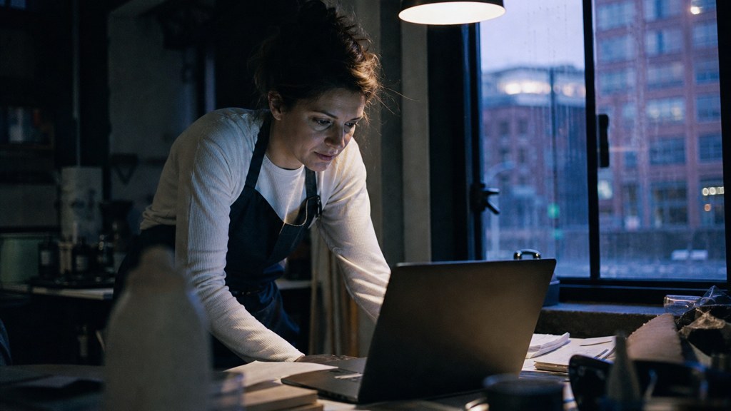 A small-business owner in an apron leaning over a laptop on a cluttered back-office desk late in the day, photographed in Manchester