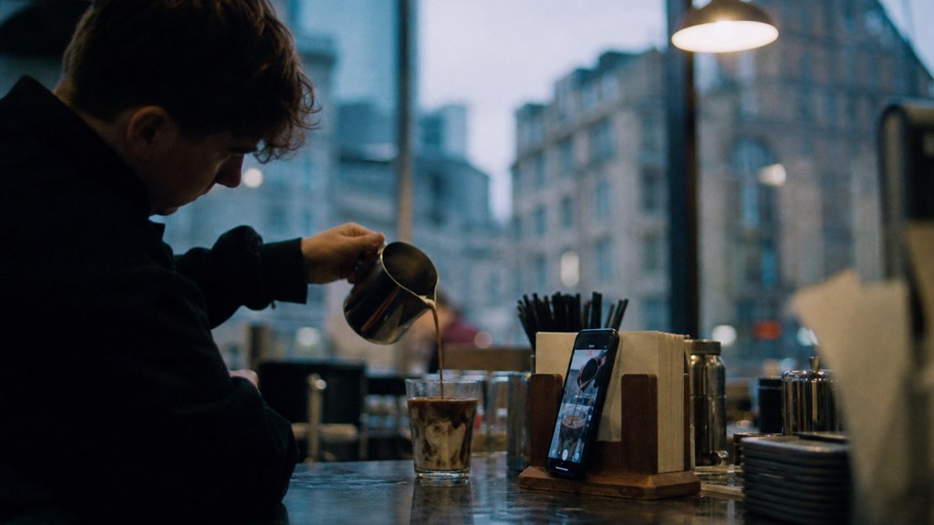 A young cafe worker filming a drink being poured on a phone propped against a napkin holder, photographed in Manchester