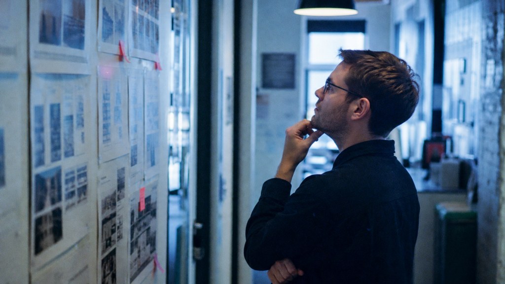 A media planner standing at a wall of printed layouts in a narrow agency corridor, photographed in Manchester