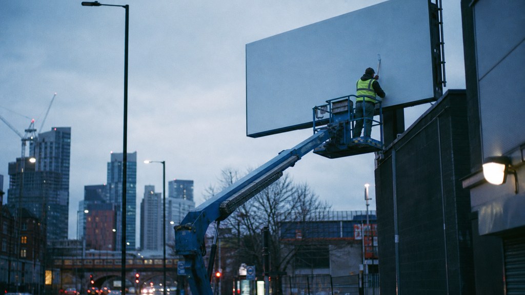 A crew member on a lift installing a blank billboard panel above a busy street, photographed in Manchester