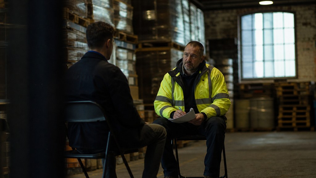 A warehouse supervisor interviewing a candidate on folding chairs beside stacked pallets, photographed in Manchester