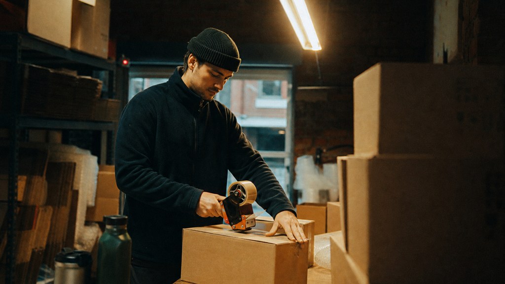 A warehouse worker taping shipping boxes under strip lighting in a small fulfilment room, photographed in Manchester