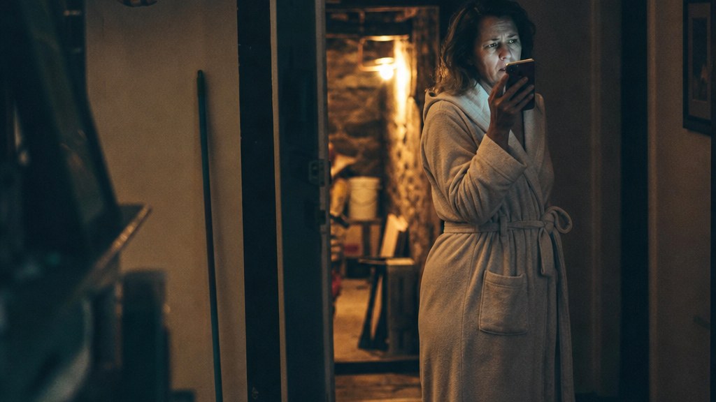 Homeowner in a dim hallway holding a phone, torchlight spilling from a basement doorway