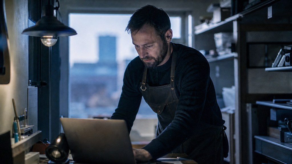 A small-business owner in an apron leaning over a laptop on a cluttered back-office desk late in the day, photographed in Sheffield