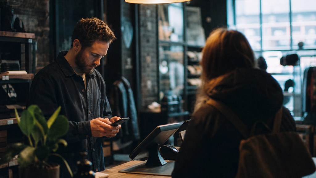A shop manager standing behind the counter checking a phone while a customer waits at the till, photographed in Sheffield