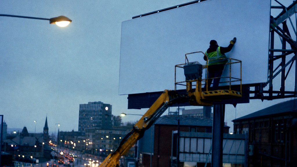 A crew member on a lift installing a blank billboard panel above a busy street, photographed in Sheffield