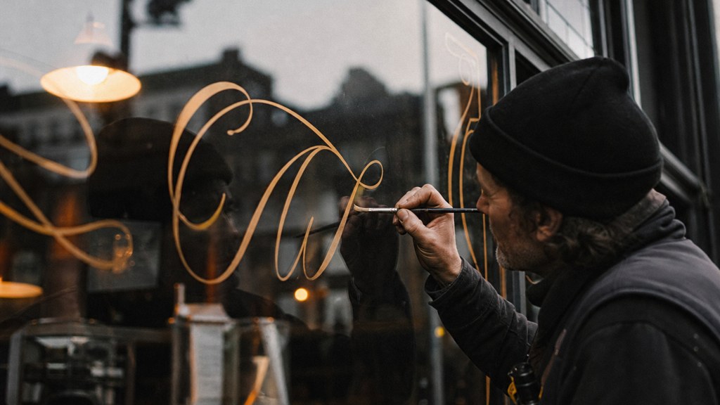 A sign painter finishing gold lettering on a shopfront window from the street side, photographed in Sheffield