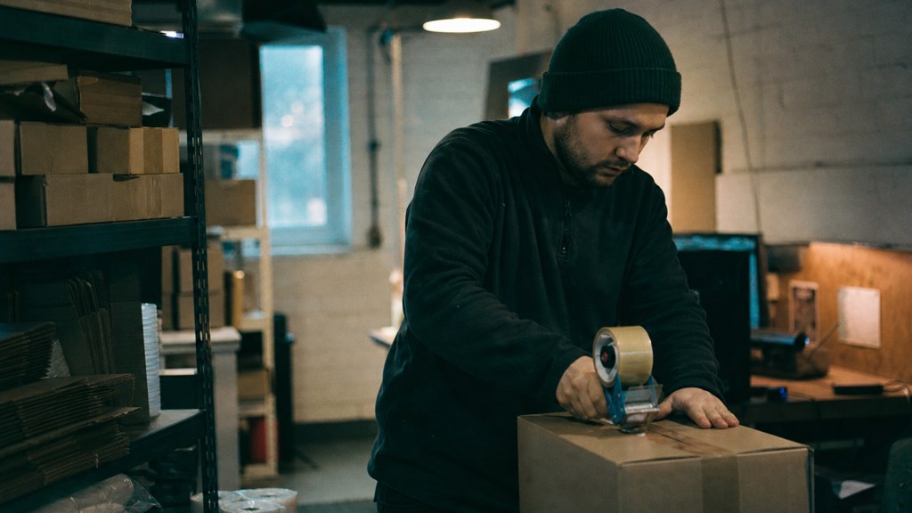 A warehouse worker taping shipping boxes under strip lighting in a small fulfilment room, photographed in Sheffield