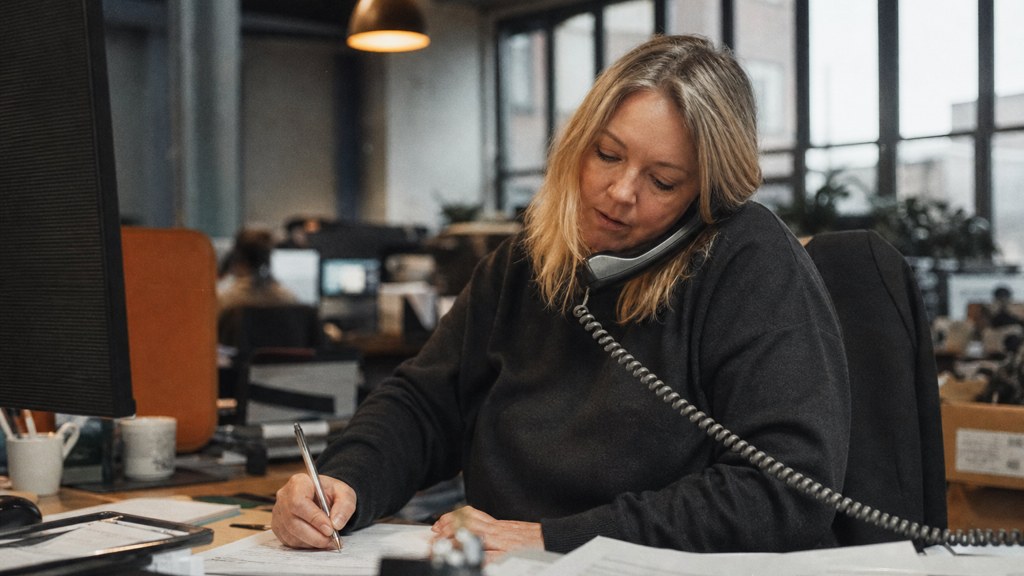 An office manager at a cluttered desk with a landline handset tucked under one ear, photographed in Sheffield