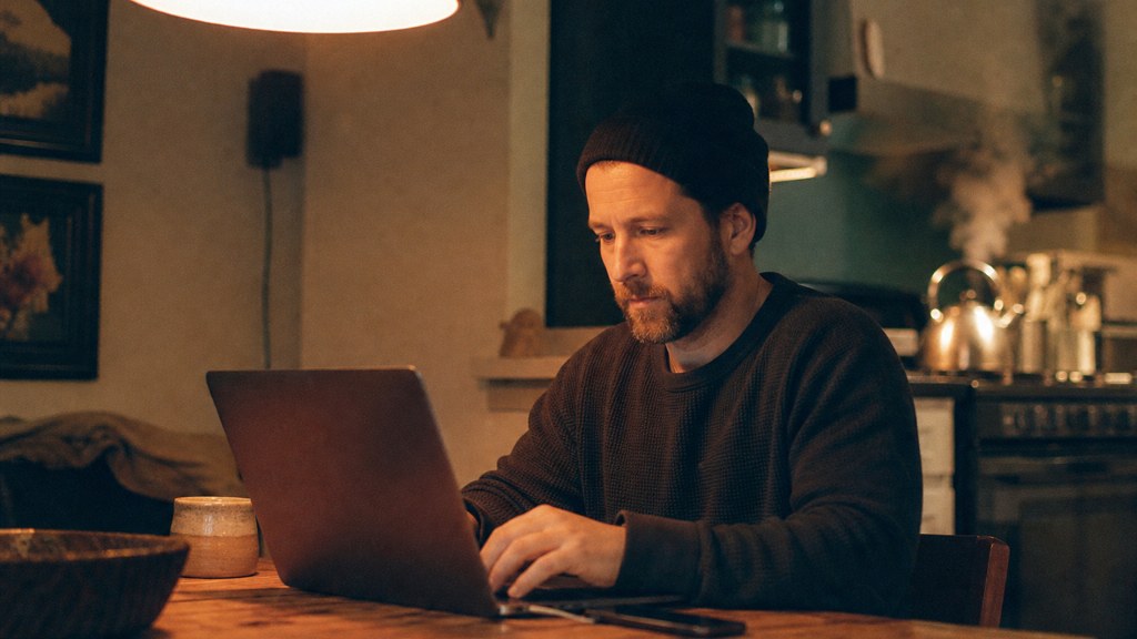 A service-business owner at a kitchen table typing a question into a laptop while a kettle steams behind, photographed in Cincinnati