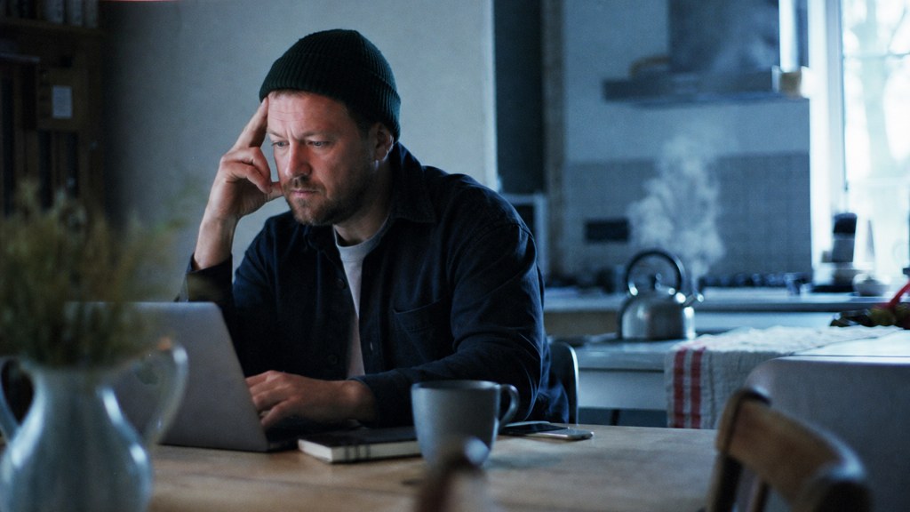 A service-business owner at a kitchen table typing a question into a laptop while a kettle steams behind, photographed in Garland